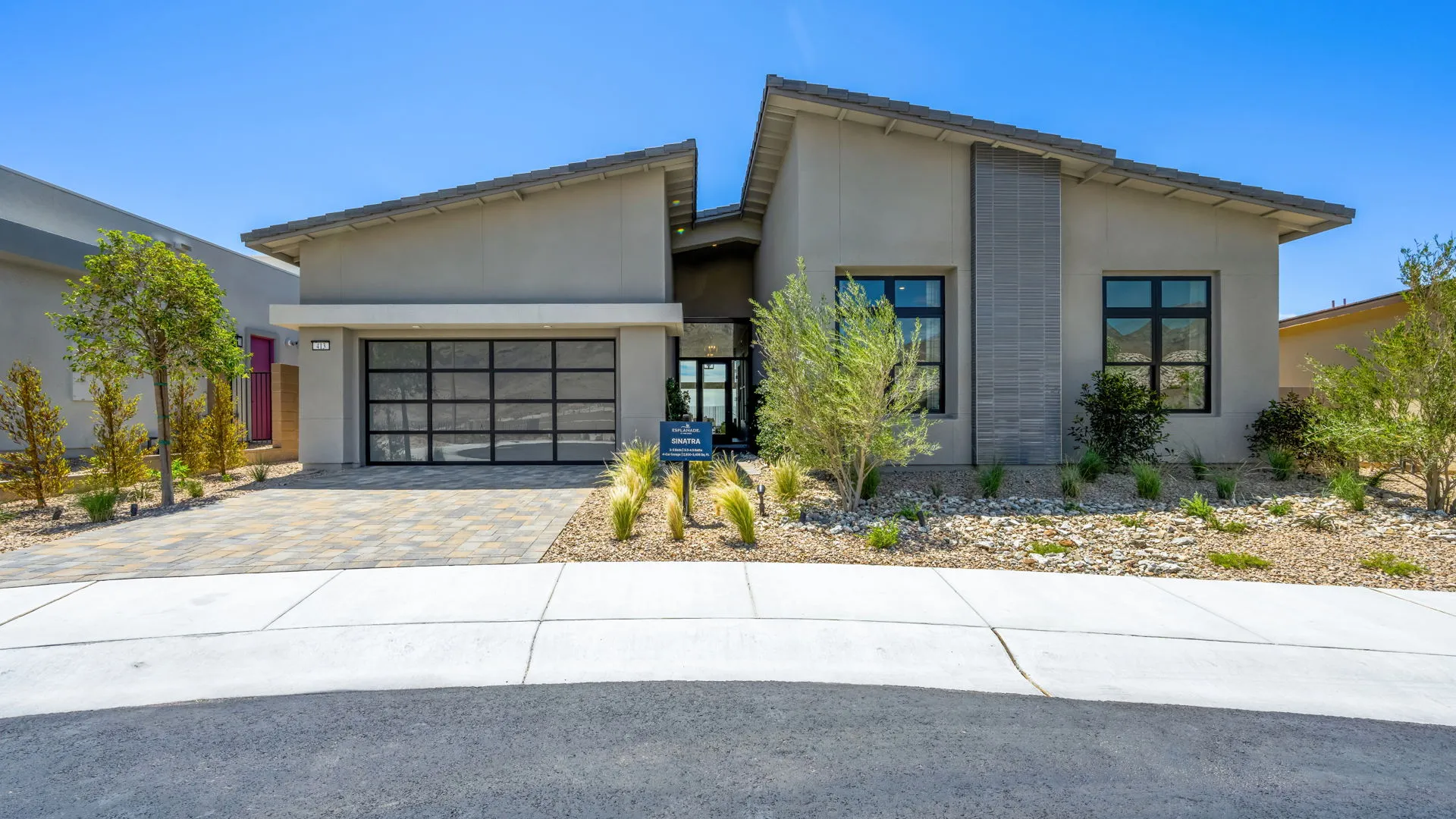 Modern single-story house with large windows, sloped roof, and glass garage door. Front yard features desert landscaping with rocks, shrubs, and small trees under a clear blue sky.