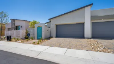 Modern single-story house with a gray garage, brick driveway, and a turquoise front gate. Minimal landscaping with rocks and small plants line the yard under a clear blue sky.