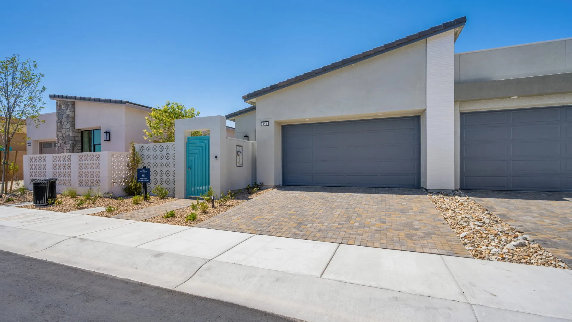Front Elevation of Tiki Model at Esplanade at Red Rock by Taylor Morrison (1) Modern single-story house with a gray garage, brick driveway, and a turquoise front gate. Minimal landscaping with rocks and small plants line the yard under a clear blue sky.