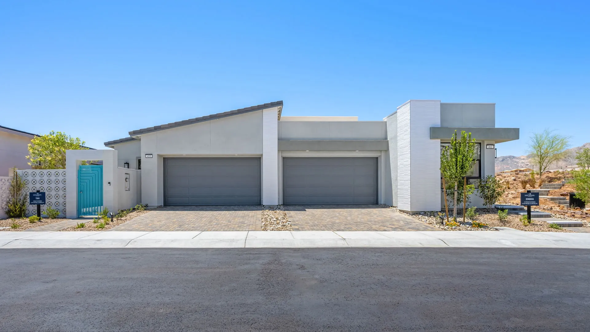 Front Elevation of Tiki Model at Esplanade at Red Rock by Taylor Morrison (2) Modern single-story house with flat roof, three gray garage doors, light-colored exterior, stone driveway, minimal landscaping, and a bright blue gate on the left under a clear blue sky.