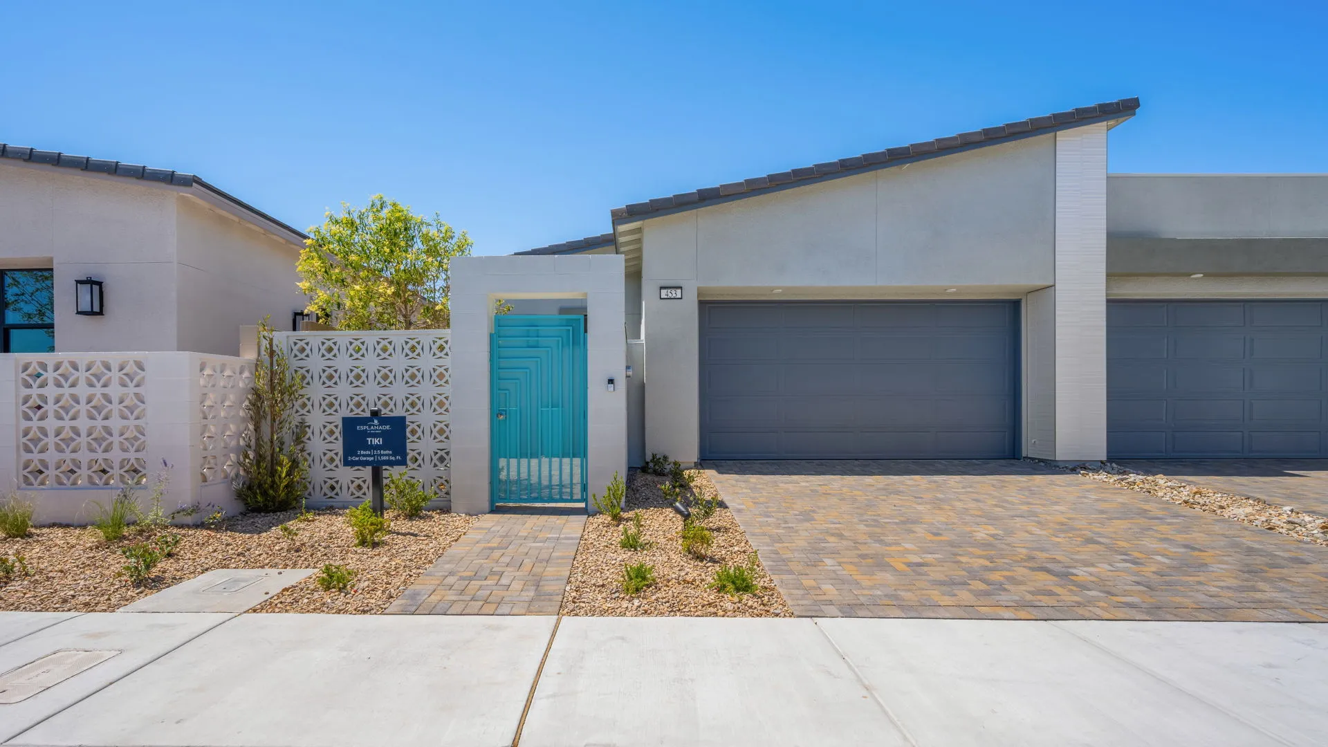 Front Elevation of Tiki Model at Esplanade at Red Rock by Taylor Morrison Modern single-story house with a light blue front gate, decorative white privacy wall, two-car garage, brick driveway, and desert landscaping under a clear blue sky.