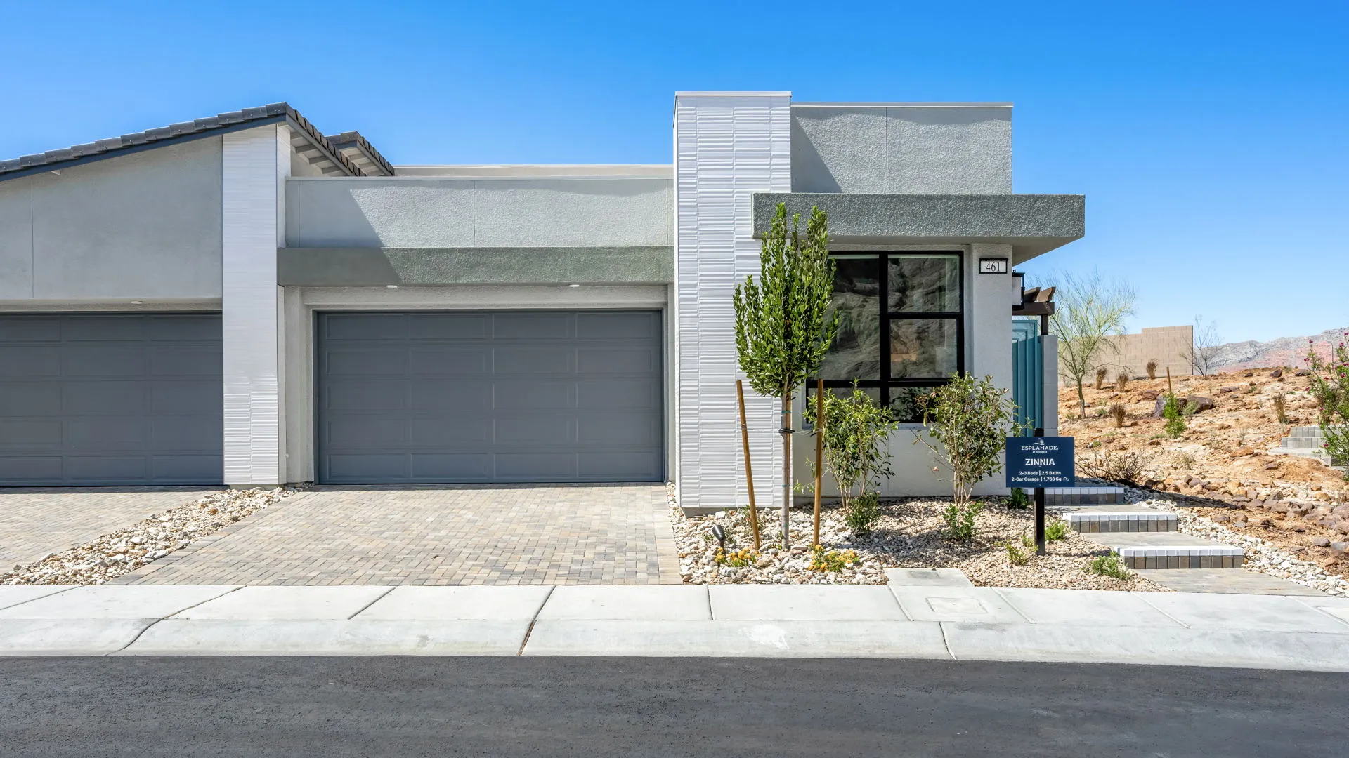 Front Elevation of Zinnia Model at Esplanade at Red Rock by Taylor Morrison (1) Modern single-story house with a flat roof, double garage, gray and white exterior, large front window, minimal desert landscaping, and a paved driveway under a clear blue sky.