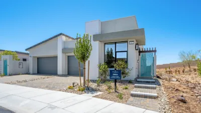 Modern single-story house with flat roof, large windows, and a three-car garage. Desert landscaping with rocks and young trees surrounds the paved walkway leading to the front door under a clear blue sky.