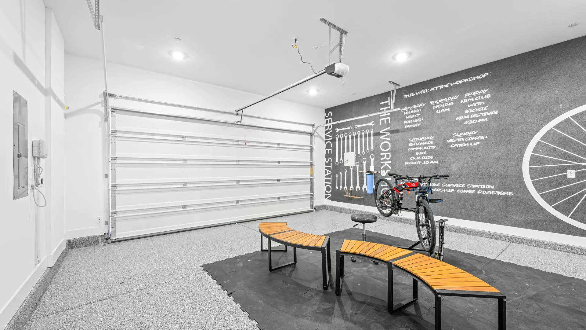 Garage of Monroe Model at Esplanade at Red Rock by Taylor Morrison (1) Modern garage with a large door, bike repair station with tools, a bike on a stand, two curved wooden benches, wall art featuring a bike wheel, and a black mat on the floor.
