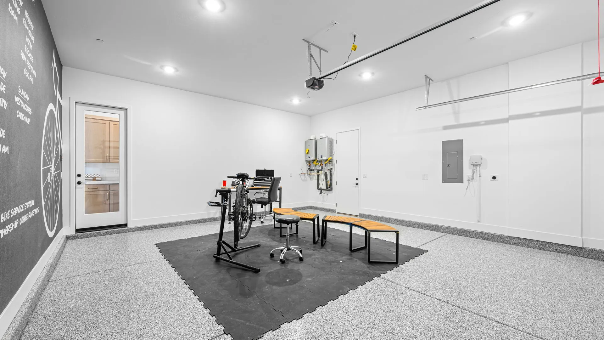 Garage of Monroe Model at Esplanade at Red Rock by Taylor Morrison A clean, modern garage with speckled flooring, white walls, a chalkboard wall, bike trainer, benches, and utility panels. Overhead lights brighten the organized, multipurpose space.