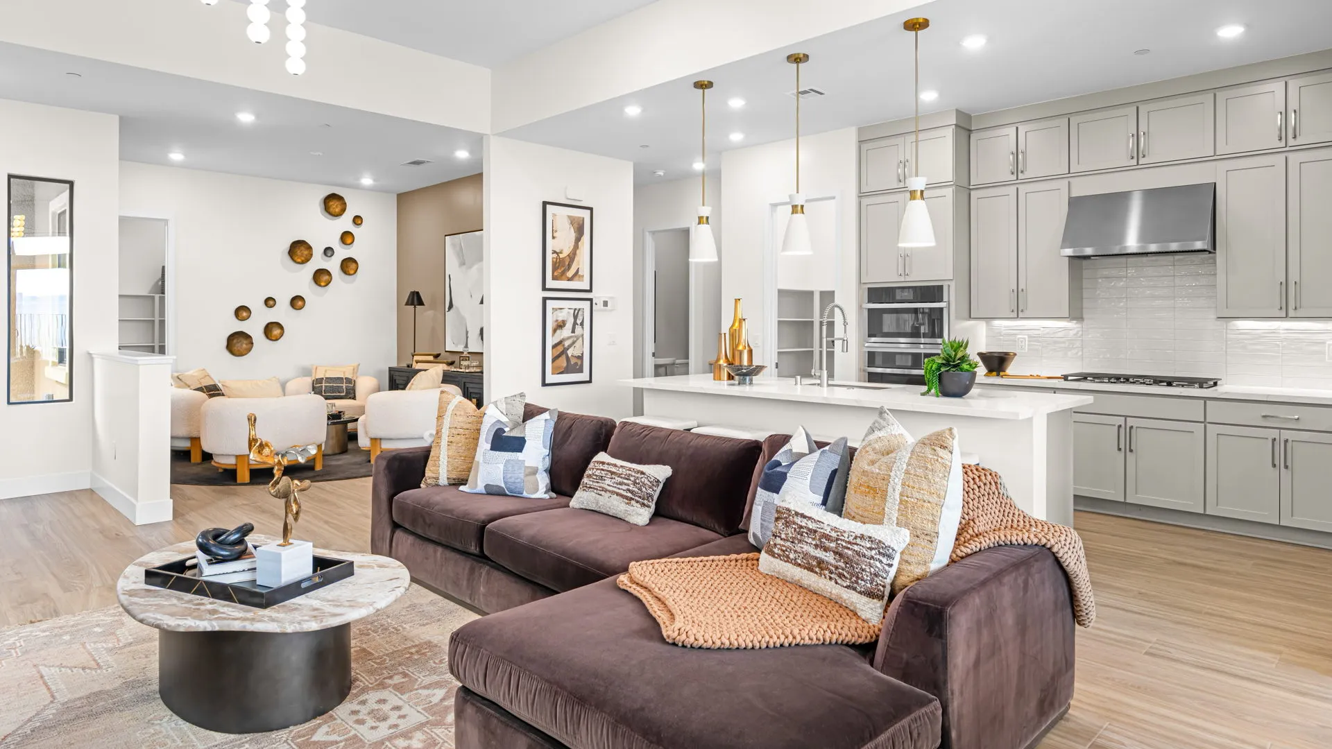 Great Room of Paloma Model at Esplanade at Red Rock by Taylor Morrison (1) Modern open-concept living room and kitchen with brown sectional sofa, decorative pillows, marble coffee table, white island with bar stools, pendant lights, and neutral-toned cabinetry.