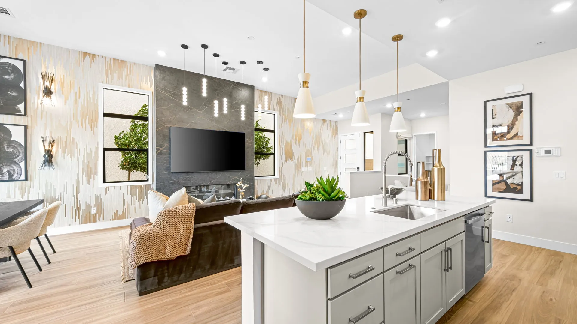 Great Room of Paloma Model at Esplanade at Red Rock by Taylor Morrison (2) Modern open-concept living space with a kitchen island, pendant lights, a sofa, wall-mounted TV, and stylish decor. Dining area and framed artwork are visible in the background.