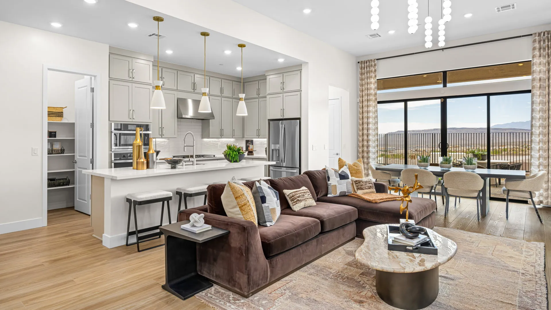 Great Room of Paloma Model at Esplanade at Red Rock by Taylor Morrison Modern open-concept living space with a brown sectional sofa, marble coffee table, stylish kitchen with pendant lights, and a dining area by large windows overlooking a scenic outdoor view.