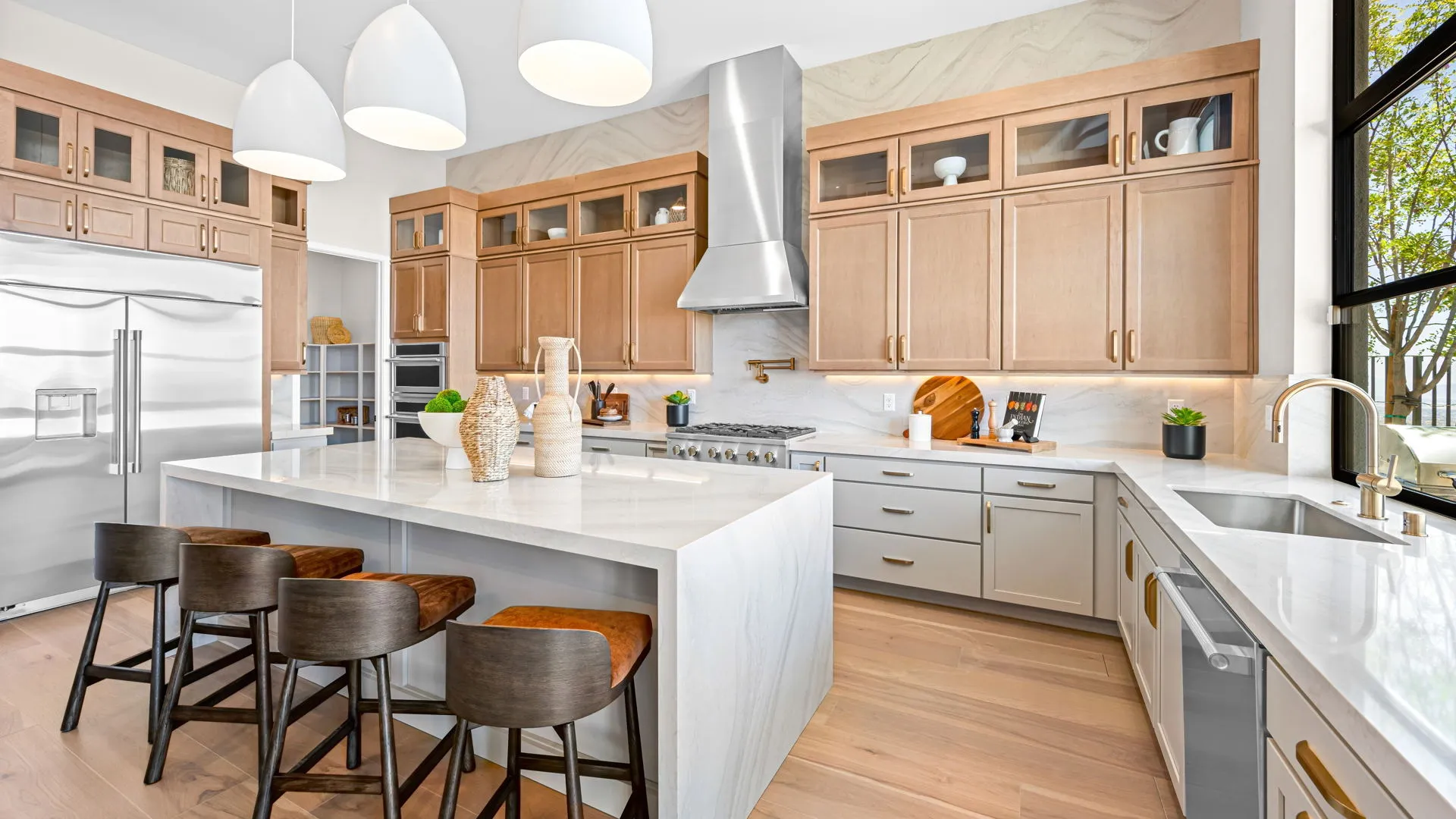 Kitchen of Monroe Model at Esplanade at Red Rock by Taylor Morrison (2) Modern kitchen with light wood cabinets, marble countertops, stainless steel appliances, pendant lights, an island with four dark chairs, and large windows providing natural light. Decor includes vases and small green plants.
