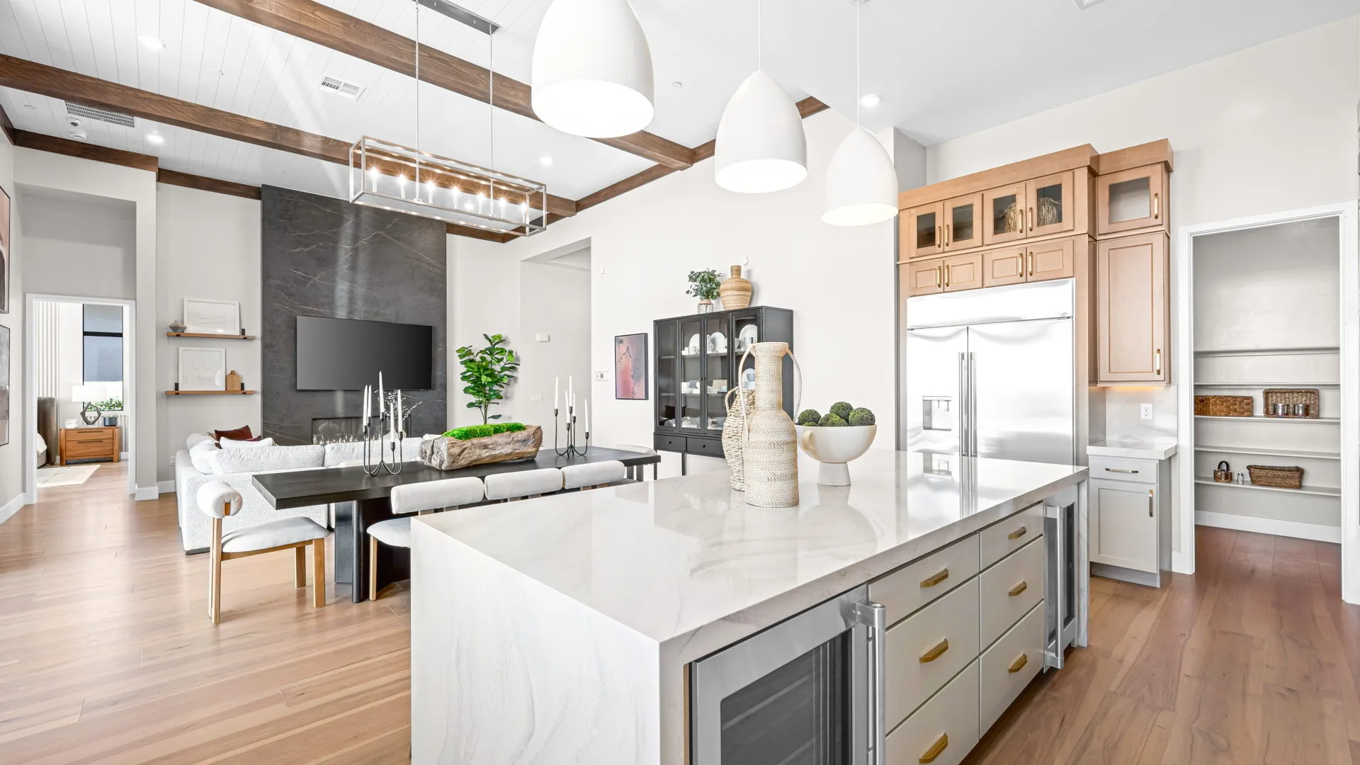 Kitchen of Monroe Model at Esplanade at Red Rock by Taylor Morrison (7) Bright, modern kitchen with a marble island, pendant lights, stainless steel appliances, and an open living area with a dining table, white chairs, and a TV on a dark accent wall. Wooden floors and ceiling beams throughout.