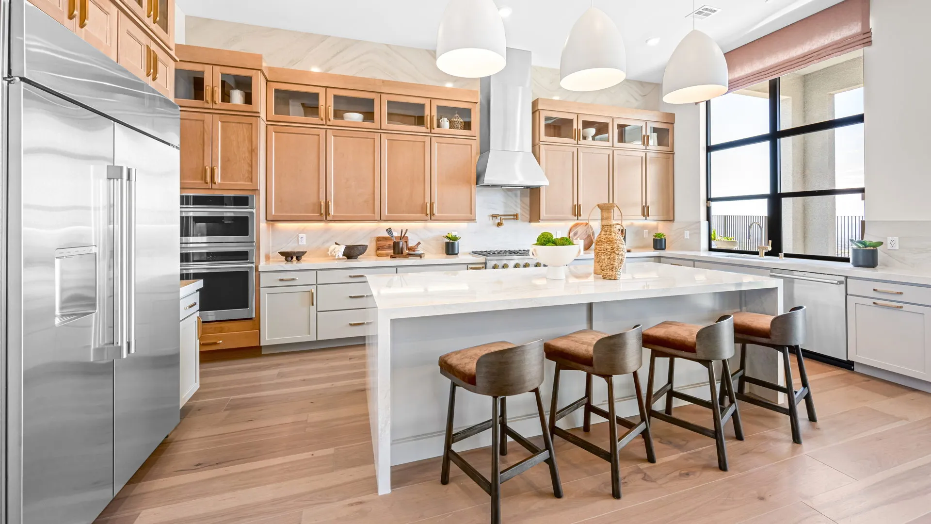 Kitchen of Monroe Model at Esplanade at Red Rock by Taylor Morrison Modern kitchen with light wood cabinets, a large island with three barstools, stainless steel appliances, pendant lights, and large windows letting in natural light.