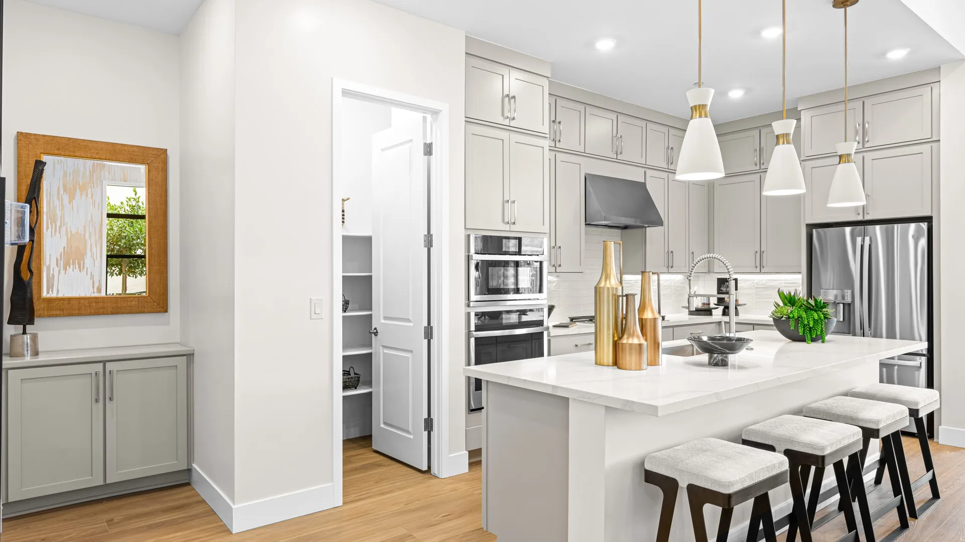 Kitchen of Paloma Model at Esplanade at Red Rock by Taylor Morrison (1) Modern kitchen with white cabinets, stainless steel appliances, a center island with three stools, pendant lights, decorative vases, and an open pantry door. Hardwood floors and a wall mirror are also visible.
