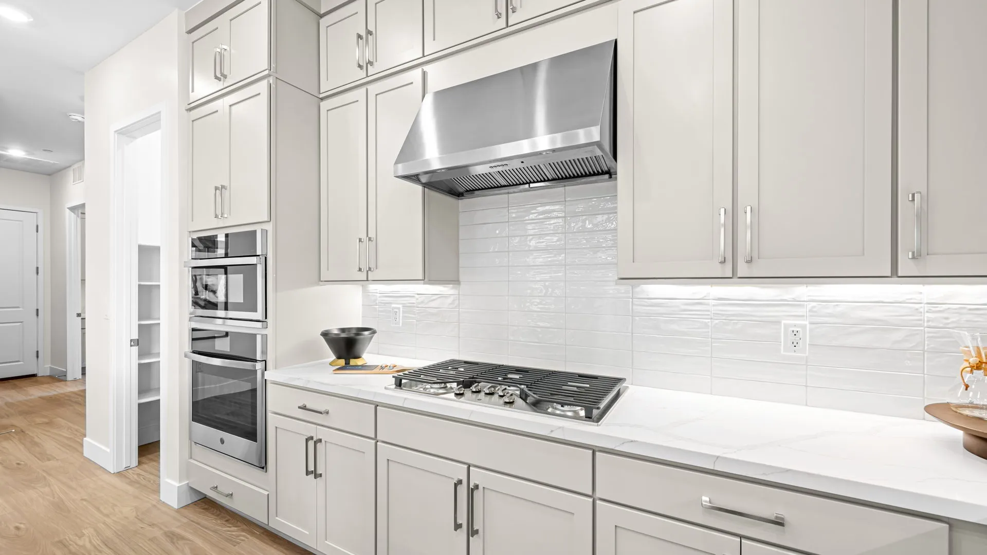 Kitchen of Paloma Model at Esplanade at Red Rock by Taylor Morrison (2) Modern kitchen with light-colored cabinets, stainless steel range hood and stove, double ovens, white tile backsplash, and wood flooring. A black bowl and a decorative item sit on the counters.