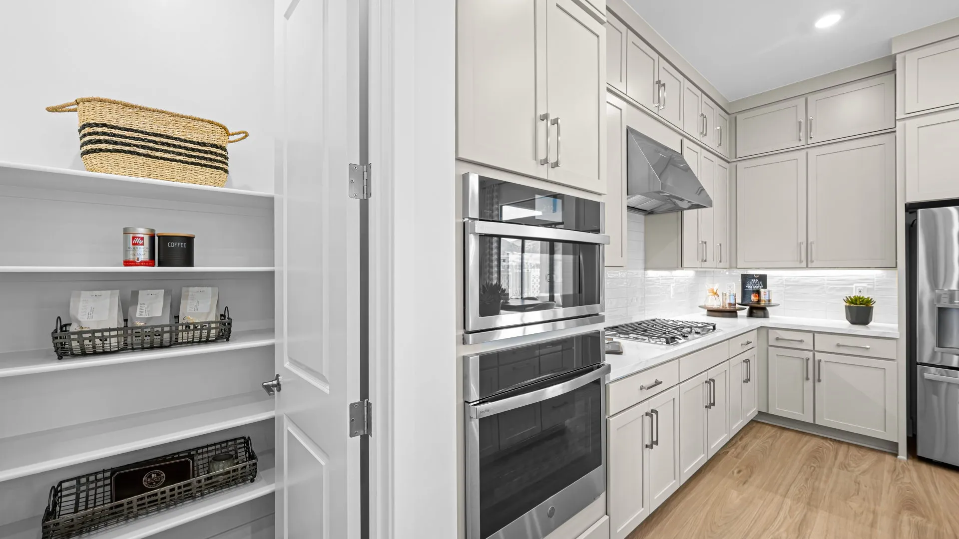 Kitchen of Paloma Model at Esplanade at Red Rock by Taylor Morrison (3) Modern kitchen with white cabinets, stainless steel appliances, a double oven, gas stove, and a walk-in pantry with shelves holding baskets and food items. Light wood flooring and bright lighting complete the space.