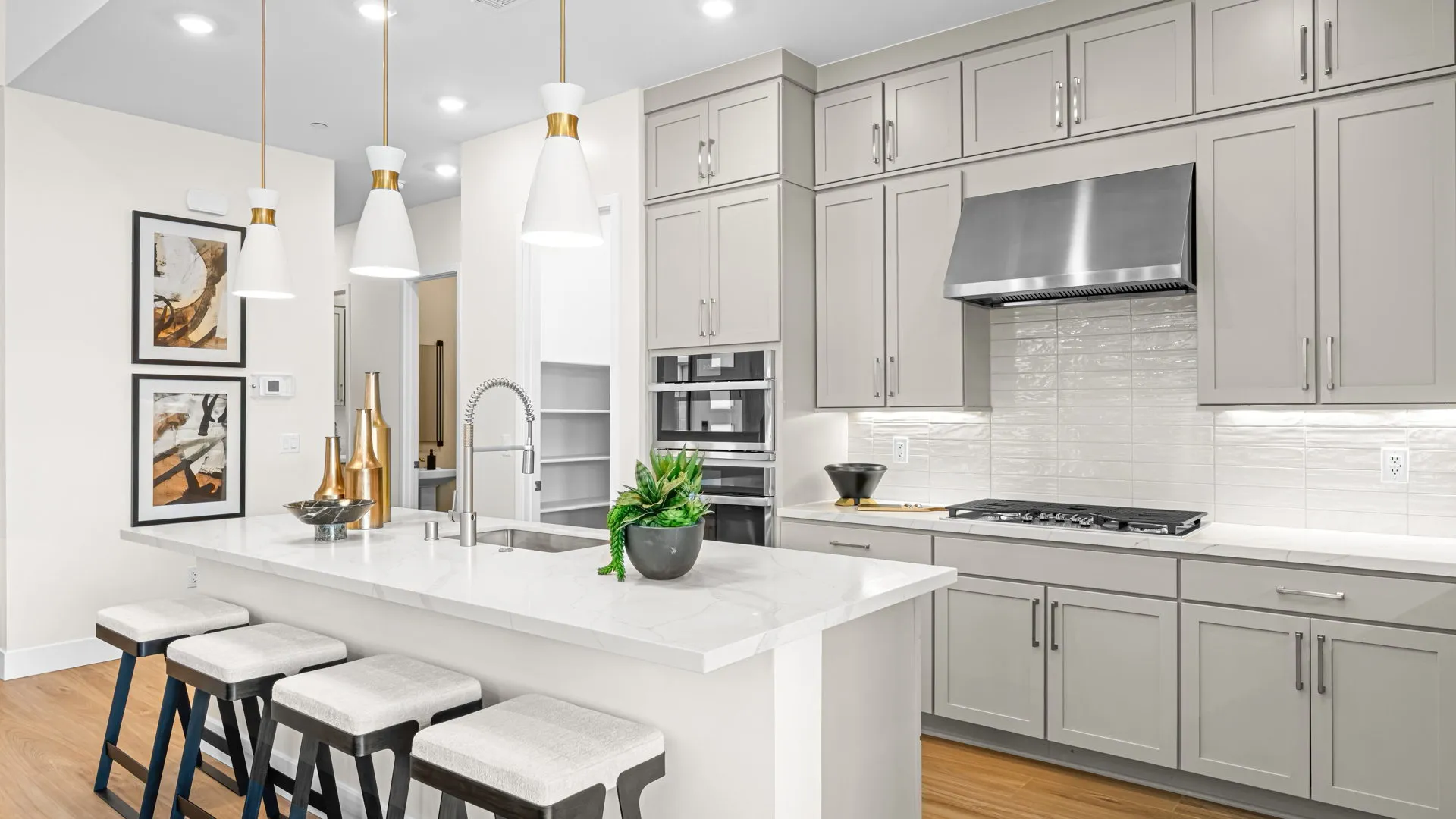 Kitchen of Paloma Model at Esplanade at Red Rock by Taylor Morrison Modern kitchen with gray cabinets, stainless steel appliances, a white island with three stools, pendant lights above, wood flooring, and decorative art on the wall. A plant and gold vases sit on the countertop.