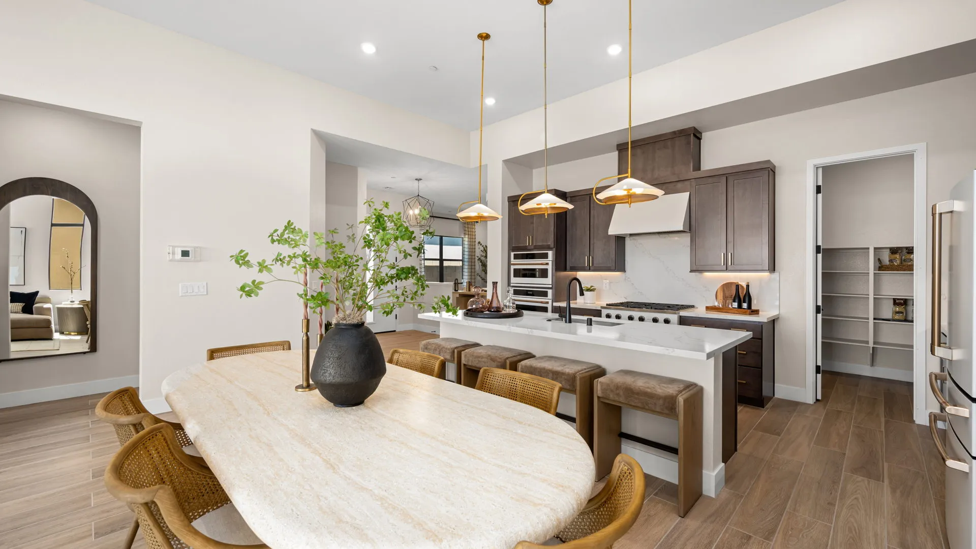 Kitchen of Sangria Model at Esplanade at Red Rock by Taylor Morrison (1) Modern kitchen with wooden cabinets, an island with barstools, a marble dining table, pendant lights, and a pantry. A vase with green branches sits on the table; the space is bright with natural light.