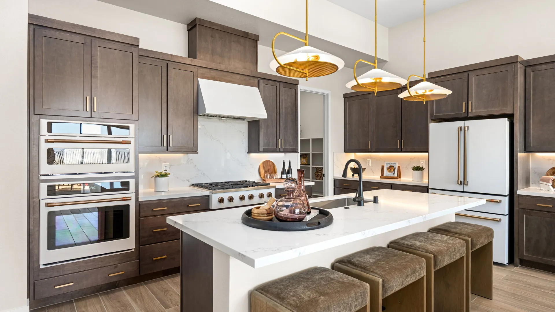 Kitchen of Sangria Model at Esplanade at Red Rock by Taylor Morrison (2) Modern kitchen with dark wood cabinets, white marble countertops, three gold pendant lights, stainless steel appliances, and upholstered stools at the island. Decorative items and a sink are on the island.