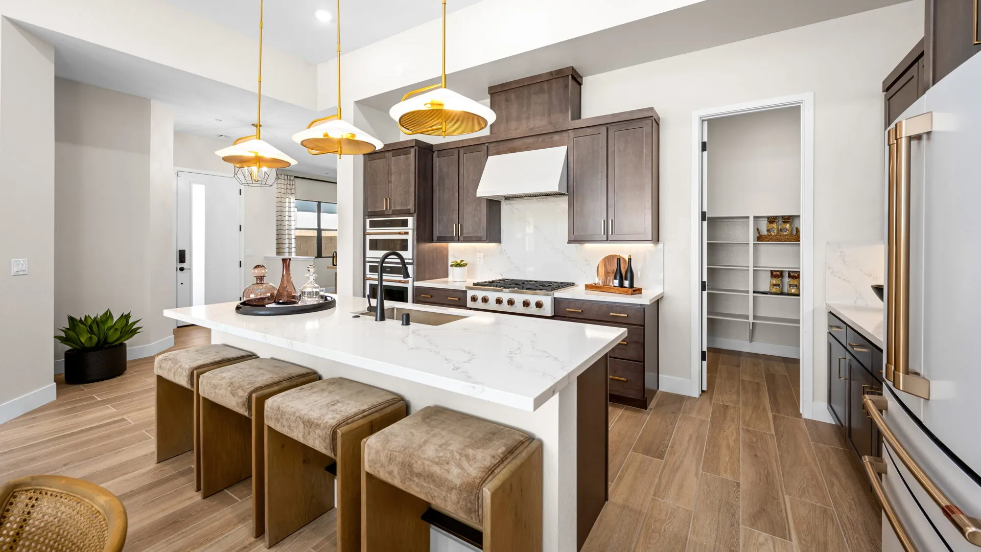 Kitchen of Sangria Model at Esplanade at Red Rock by Taylor Morrison (3) Modern kitchen with dark wood cabinets, white countertops, an island with three cushioned stools, gold pendant lights, and an open pantry with shelves partially stocked with food items. Light wood floors throughout.