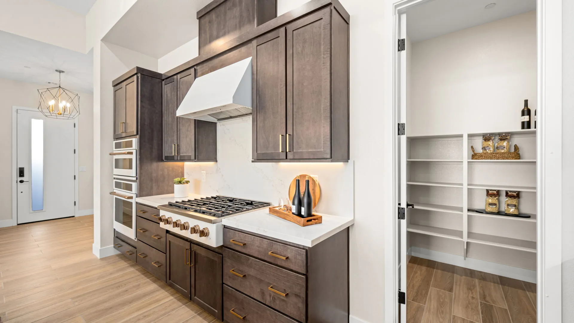 Kitchen of Sangria Model at Esplanade at Red Rock by Taylor Morrison (4) Modern kitchen with dark cabinets, white countertops, stainless steel appliances, a gas stove, and open pantry shelves with food items. The floor is light wood, and the space has bright, natural lighting.