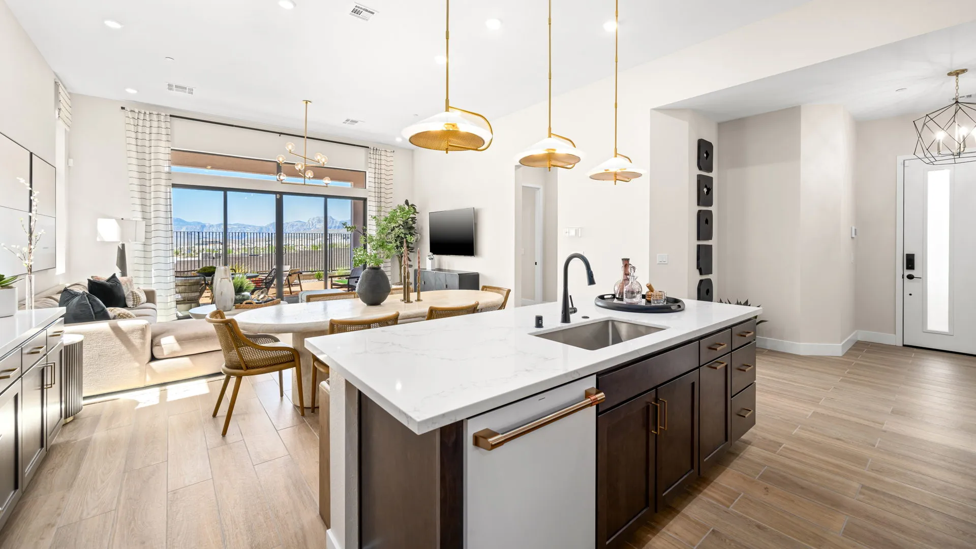 Kitchen of Sangria Model at Esplanade at Red Rock by Taylor Morrison (5) Modern open-concept kitchen and living area with white island, gold fixtures, wood floors, large windows, mountain views, dining table, and neutral decor.