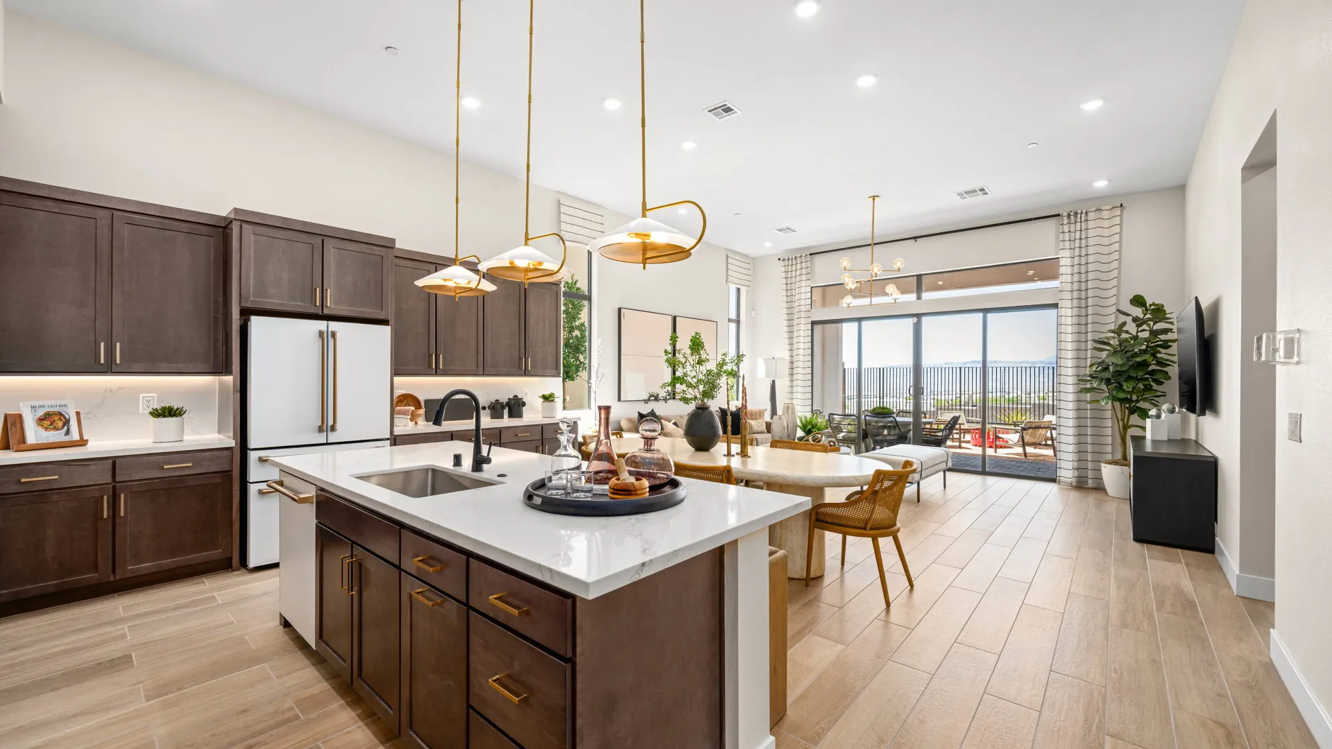 Kitchen of Sangria Model at Esplanade at Red Rock by Taylor Morrison (6) Modern kitchen with dark cabinets, white countertops, island with sink, pendant lights, and open layout leading to a bright living area with large windows and outdoor patio view.