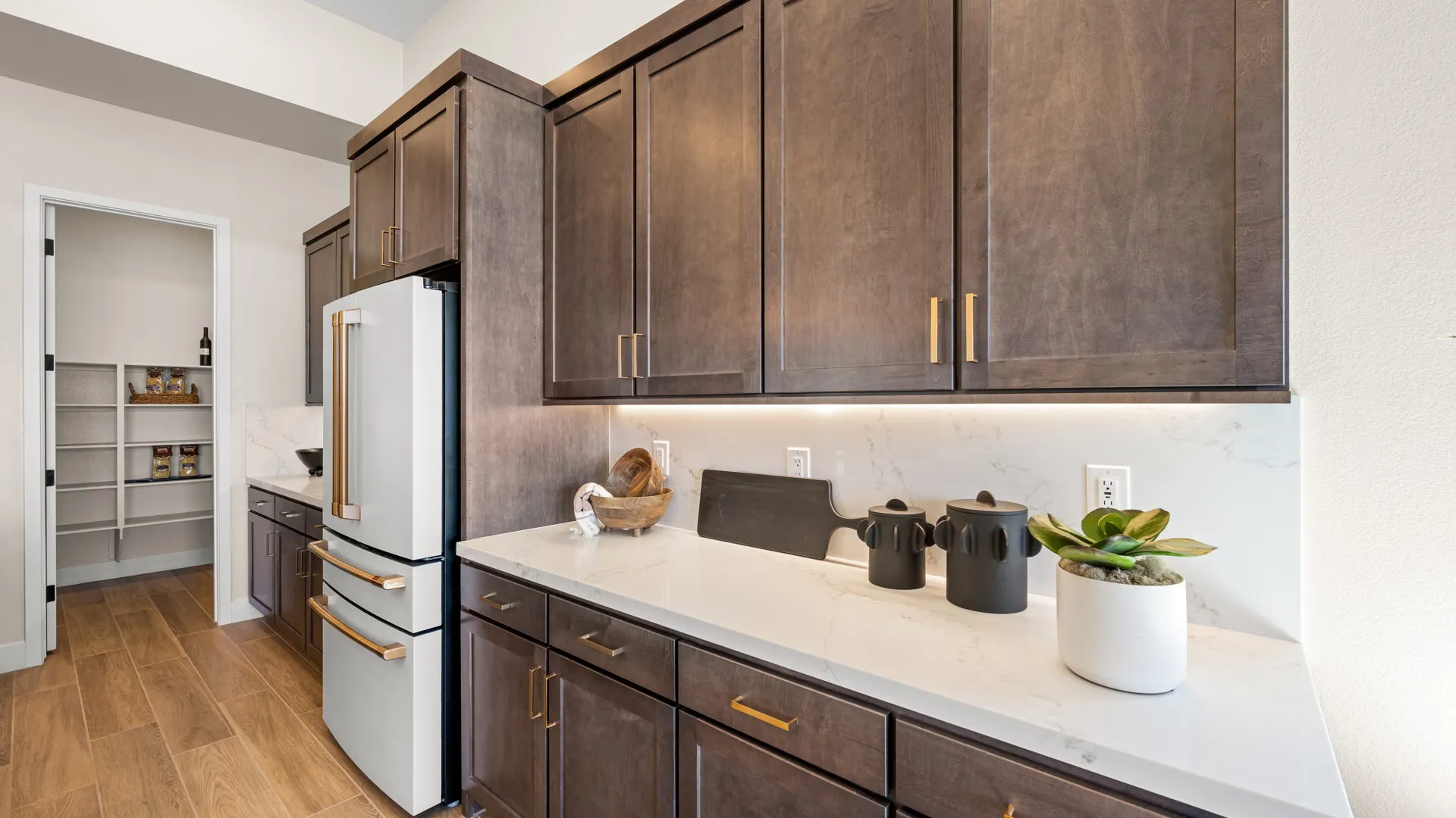 Kitchen of Sangria Model at Esplanade at Red Rock by Taylor Morrison (7) Modern kitchen with dark wood cabinets, white countertops, stainless steel refrigerator, black and white decor items, and a pantry with shelves visible through an open door.