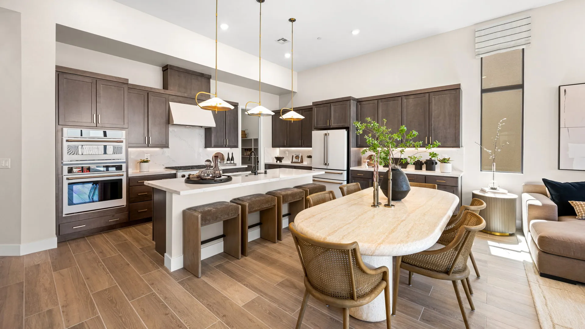 Kitchen of Sangria Model at Esplanade at Red Rock by Taylor Morrison Modern kitchen and dining area with wood cabinets, white countertops, stainless steel appliances, a large island with stools, oval dining table, woven chairs, and decorative plants, all with wood-look flooring.