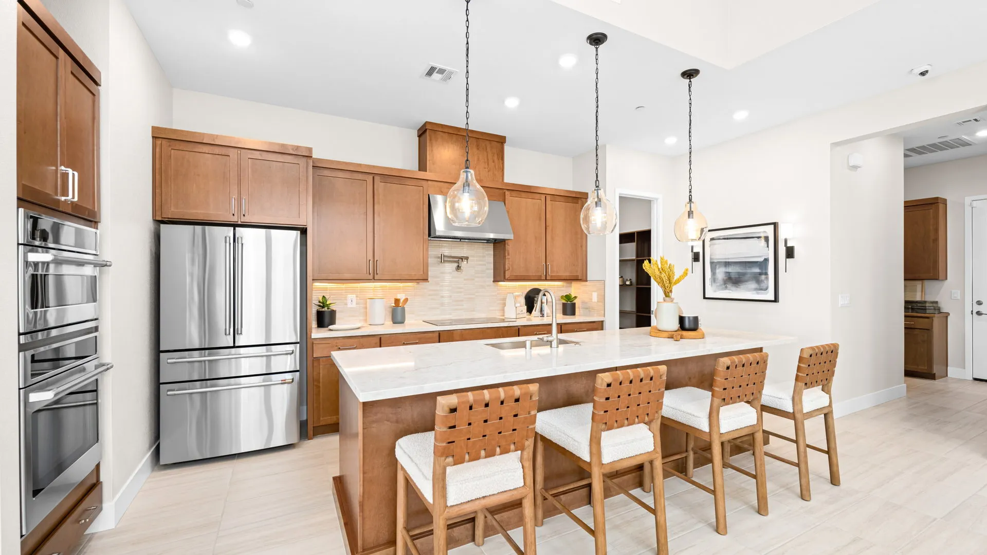 Kitchen of Tiki Model at Esplanade at Red Rock by Taylor Morrison (1) Modern kitchen with wooden cabinets, stainless steel appliances, a large island with white countertop, and four woven barstools. Pendant lights hang above the island. Decor includes plants and kitchen items.