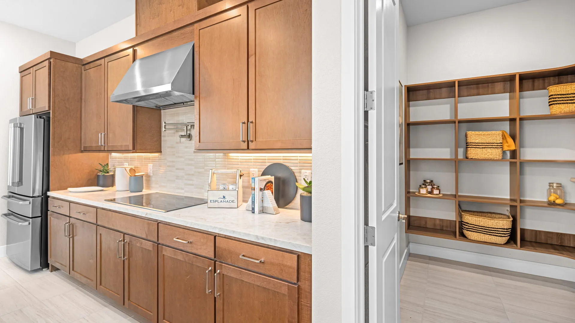 Kitchen of Tiki Model at Esplanade at Red Rock by Taylor Morrison (3) Modern kitchen with wooden cabinets, stainless steel appliances, under-cabinet lighting, light countertops, and a pantry area with shelves stocked with baskets and jars. Clean, bright, and organized with decorative accents.