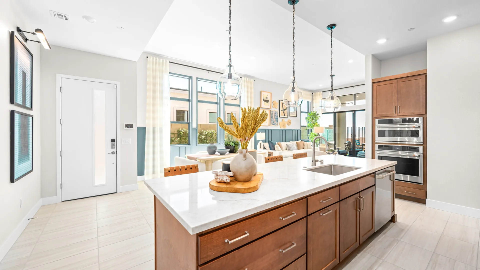 Kitchen of Tiki Model at Esplanade at Red Rock by Taylor Morrison (4) Bright modern kitchen with island, pendant lights, wooden cabinets, built-in ovens, and a sink. A vase and tray sit on the island. Large windows and an open living area are visible in the background.