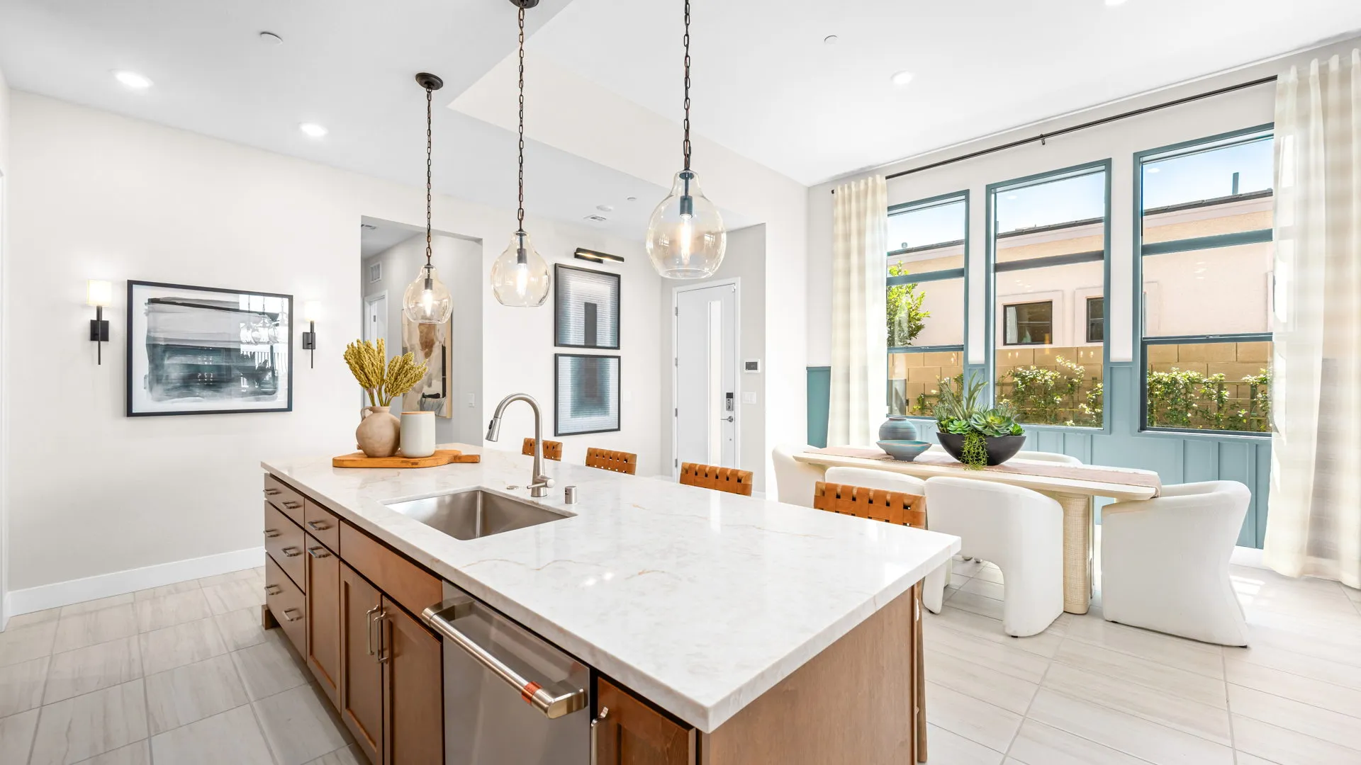 Kitchen of Tiki Model at Esplanade at Red Rock by Taylor Morrison (5) Bright modern kitchen with a marble island, brown cabinets, pendant lights, and an adjacent dining area with large windows, white chairs, a wooden table, and light curtains.