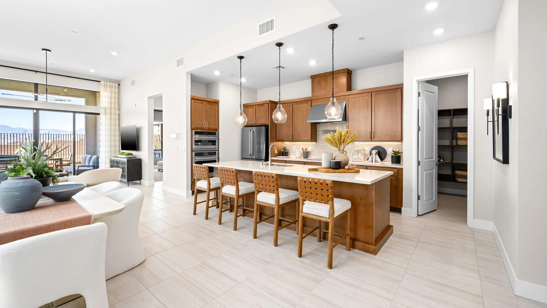 Kitchen of Tiki Model at Esplanade at Red Rock by Taylor Morrison Modern kitchen with wood cabinets, an island with four stools, pendant lights, and stainless steel appliances. Open layout leads to living and dining areas, with a pantry door open in the background.