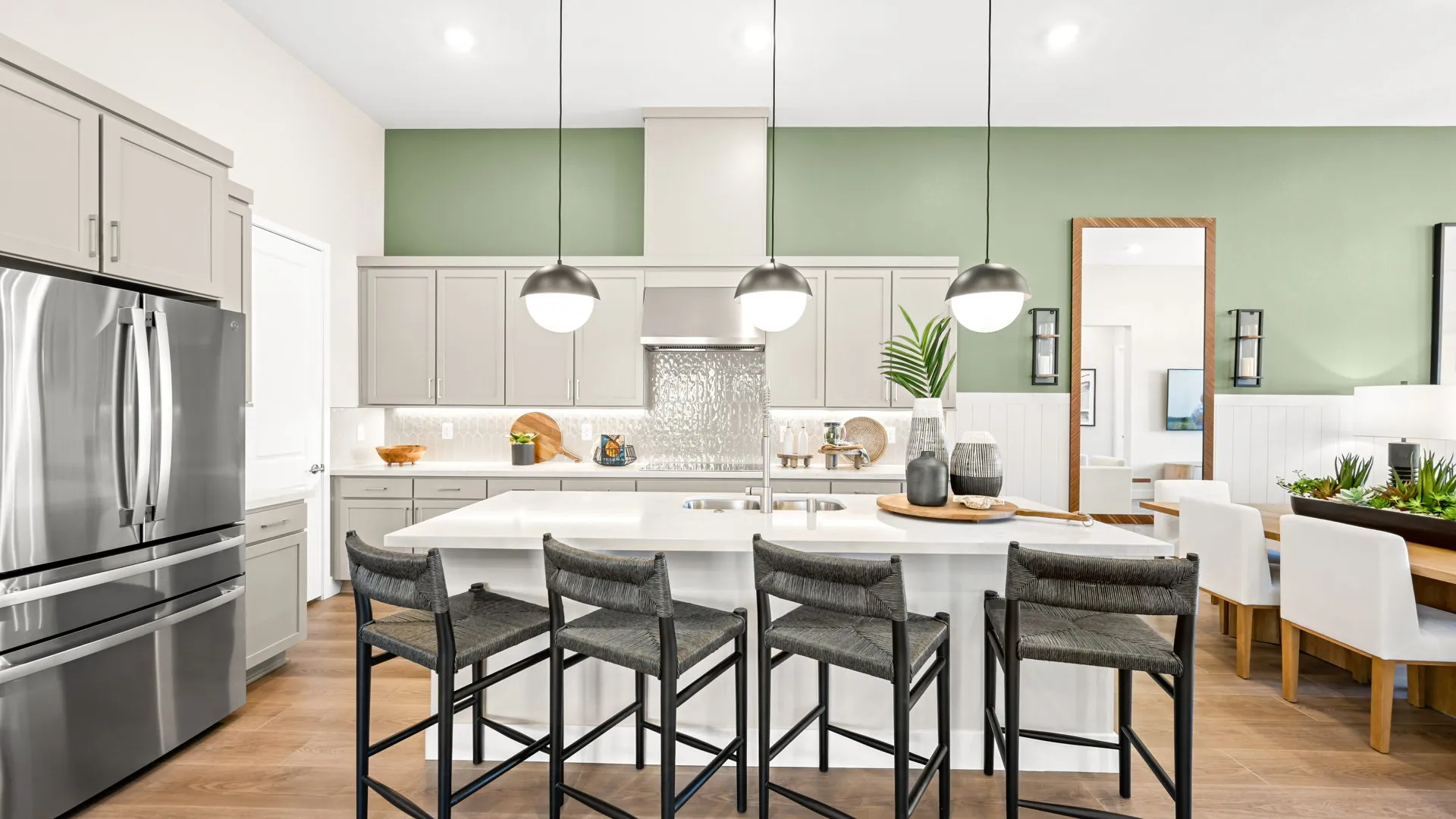 Kitchen of Zinnia Model at Esplanade at Red Rock by Taylor Morrison (1) Modern kitchen with light gray cabinets, pendant lights over a white island, four dark barstools, stainless steel appliances, and a dining area with a wooden table and white chairs in the background.