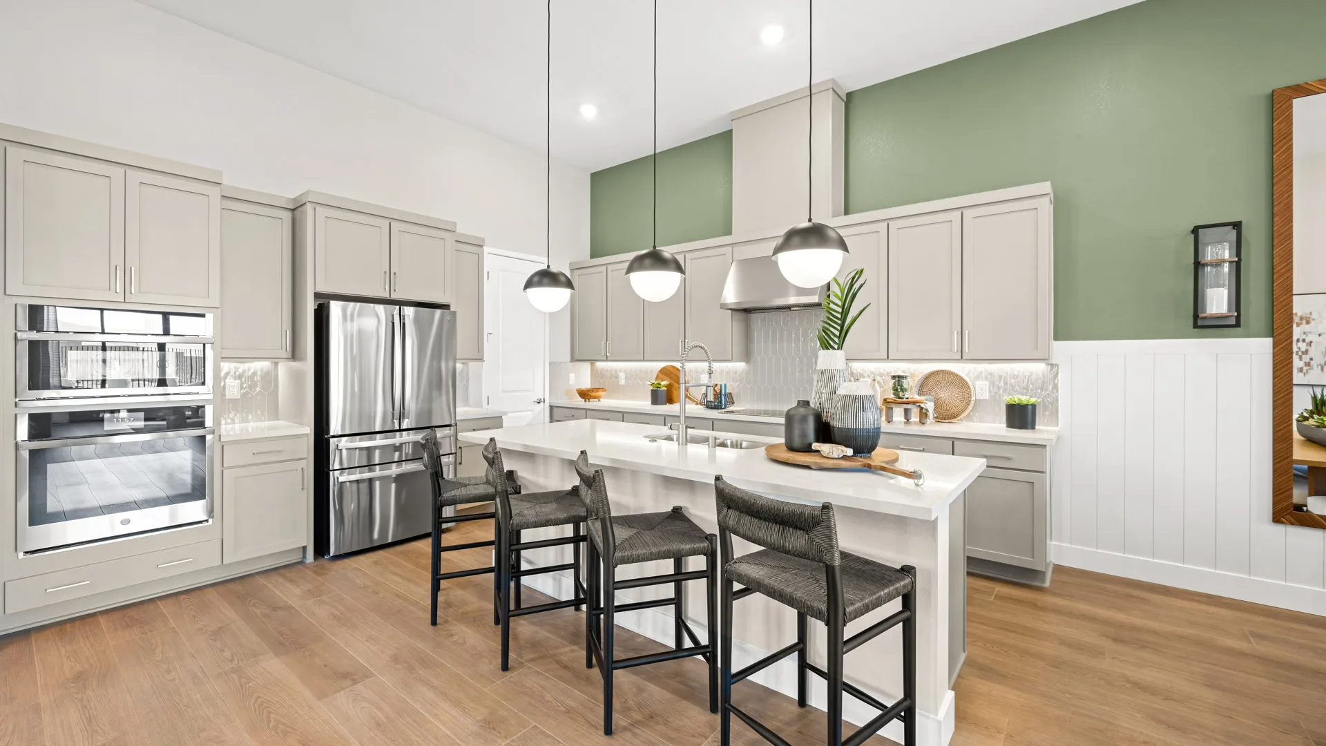 Kitchen of Zinnia Model at Esplanade at Red Rock by Taylor Morrison (2) Modern kitchen with light gray cabinets, stainless steel appliances, a green accent wall, island with white countertop, four dark chairs, pendant lights, and wood flooring. Decorative items are on the counters and shelves.