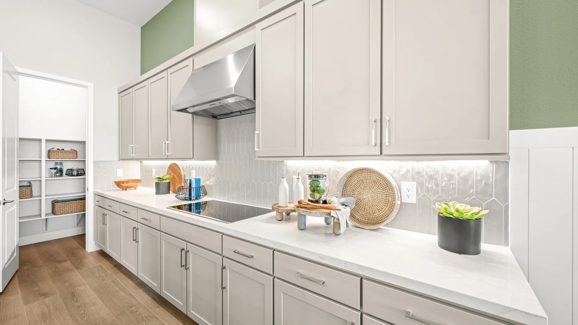 Kitchen of Zinnia Model at Esplanade at Red Rock by Taylor Morrison (3) Modern kitchen with light cabinets, a cooktop, silver vent hood, white backsplash, and white countertops. Decorative items and plants sit on the counters, and an open pantry with shelves is visible on the left.