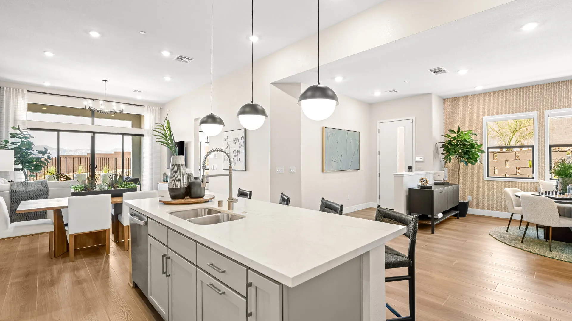 Kitchen of Zinnia Model at Esplanade at Red Rock by Taylor Morrison (4) Bright, modern open-concept kitchen and living area with a central island, pendant lights, light wood floors, dining table, plants, and large windows letting in natural light.