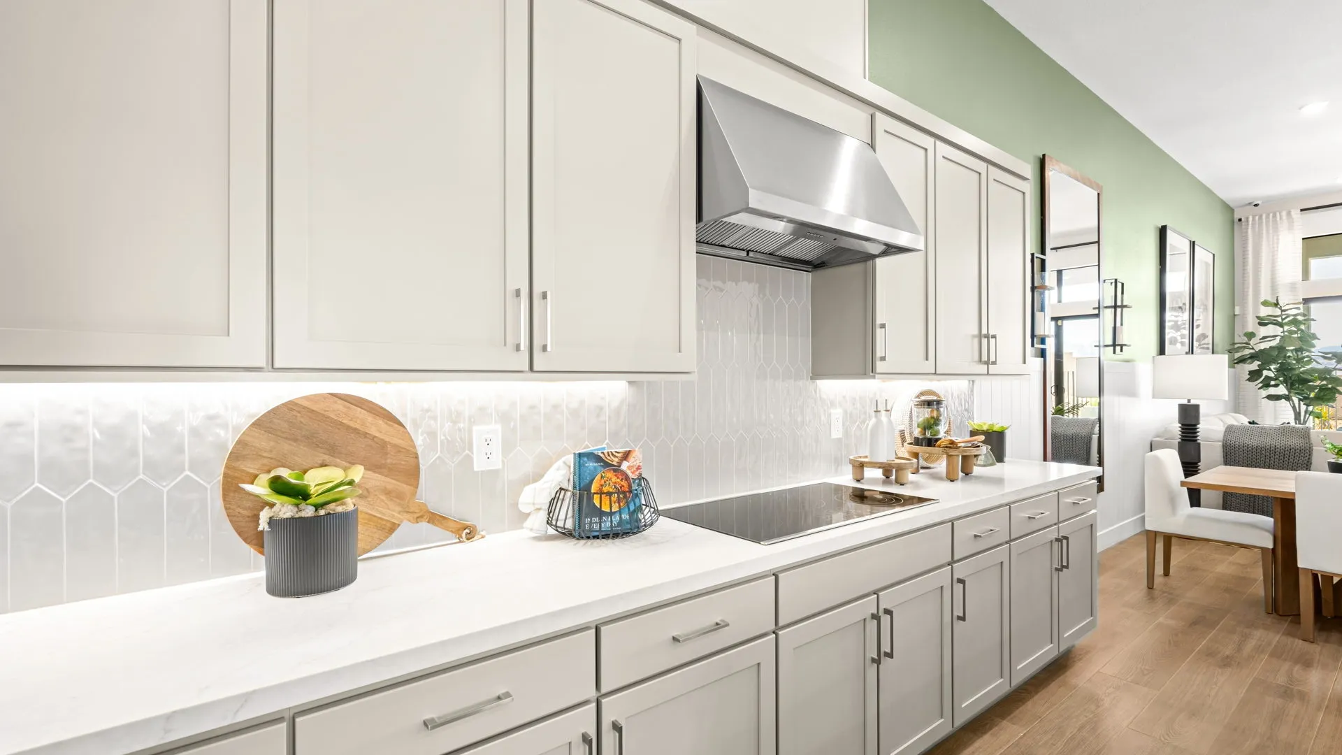 Kitchen of Zinnia Model at Esplanade at Red Rock by Taylor Morrison (5) Modern kitchen with white cabinets, a stove, stainless steel vent hood, and green accent wall. Countertop holds a cutting board, potted plant, and ceramic canisters. Dining area visible in the background.
