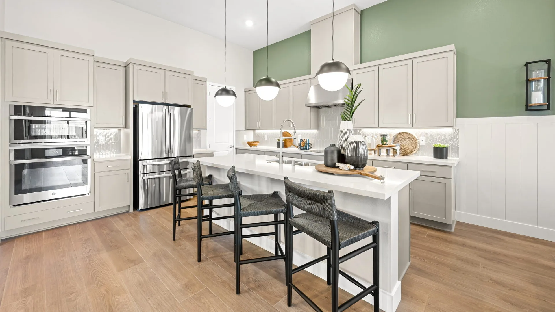 Kitchen of Zinnia Model at Esplanade at Red Rock by Taylor Morrison Modern kitchen with light gray cabinets, stainless steel appliances, a white island with three dark chairs, pendant lights above, green accent wall, and wood flooring. Decor includes plants and kitchenware.