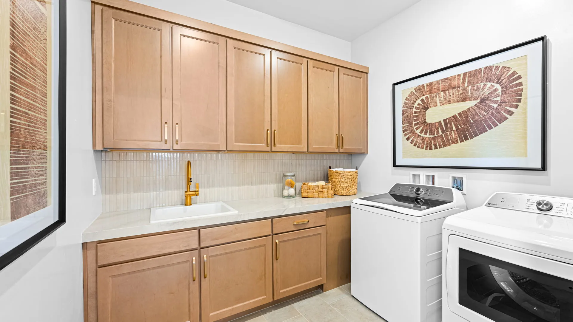 Laundry Room of Monroe Model at Esplanade at Red Rock by Taylor Morrison A modern laundry room with light wood cabinets, a white countertop, sink, gold faucet, washing machine, dryer, wicker baskets, and framed abstract art on white walls.