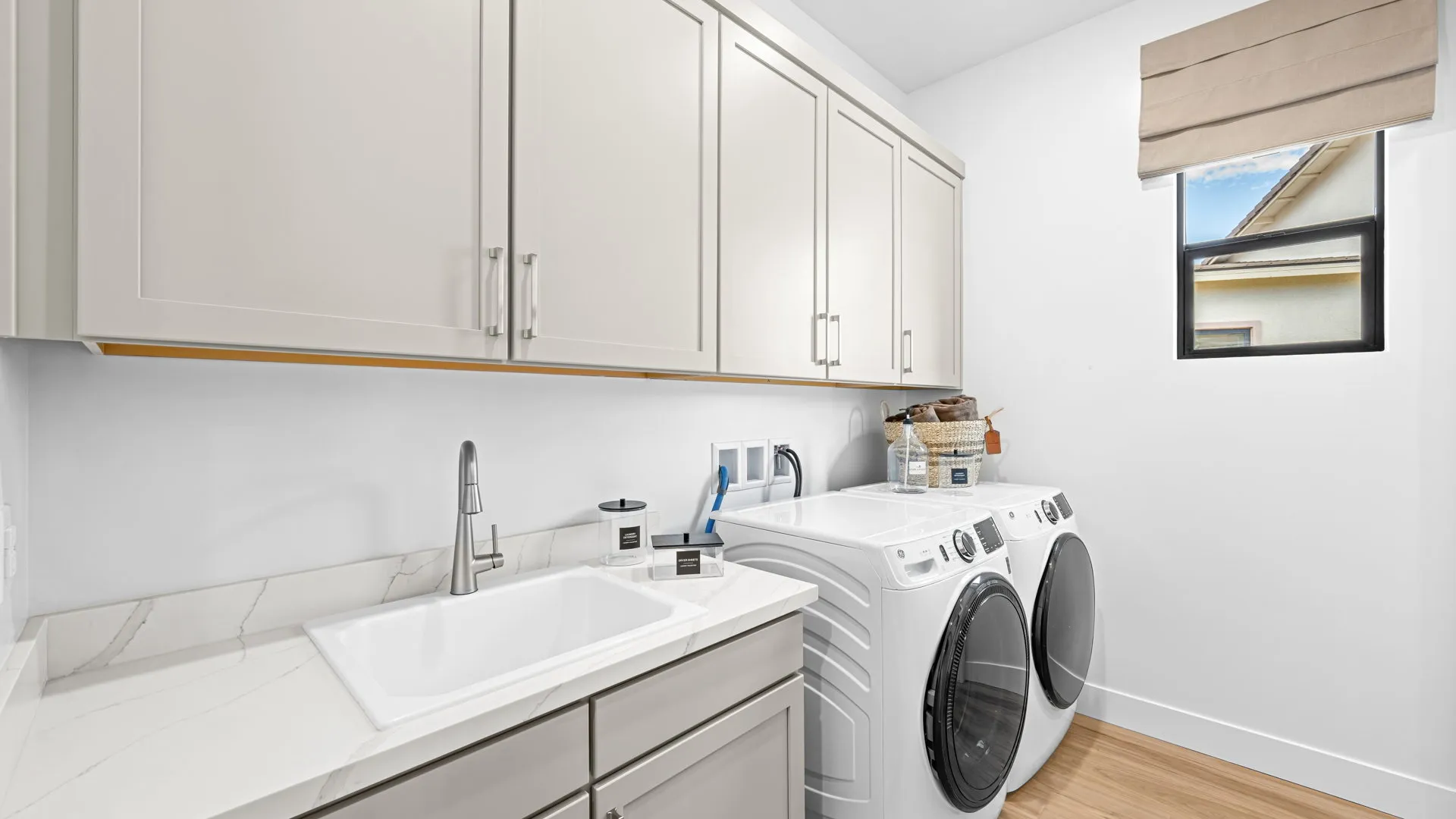 Laundry Room of Paloma Model at Esplanade at Red Rock by Taylor Morrison Modern laundry room with beige cabinets, white countertop, sink, washing machine, dryer, laundry supplies, and a window with a beige shade. Light wood floor and bright, clean atmosphere.
