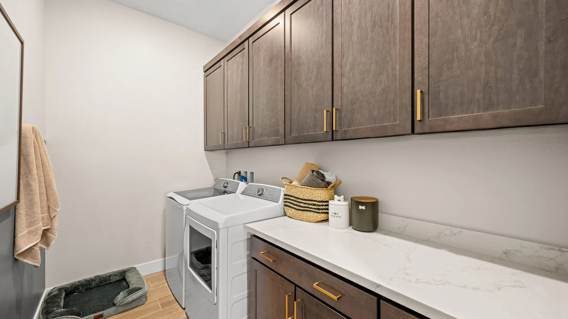 Laundry Room of Sangria Model at Esplanade at Red Rock by Taylor Morrison Modern laundry room with gray cabinets, gold handles, a washer and dryer, white countertop, laundry supplies, towels, and a small green pet bed on a light wood floor.
