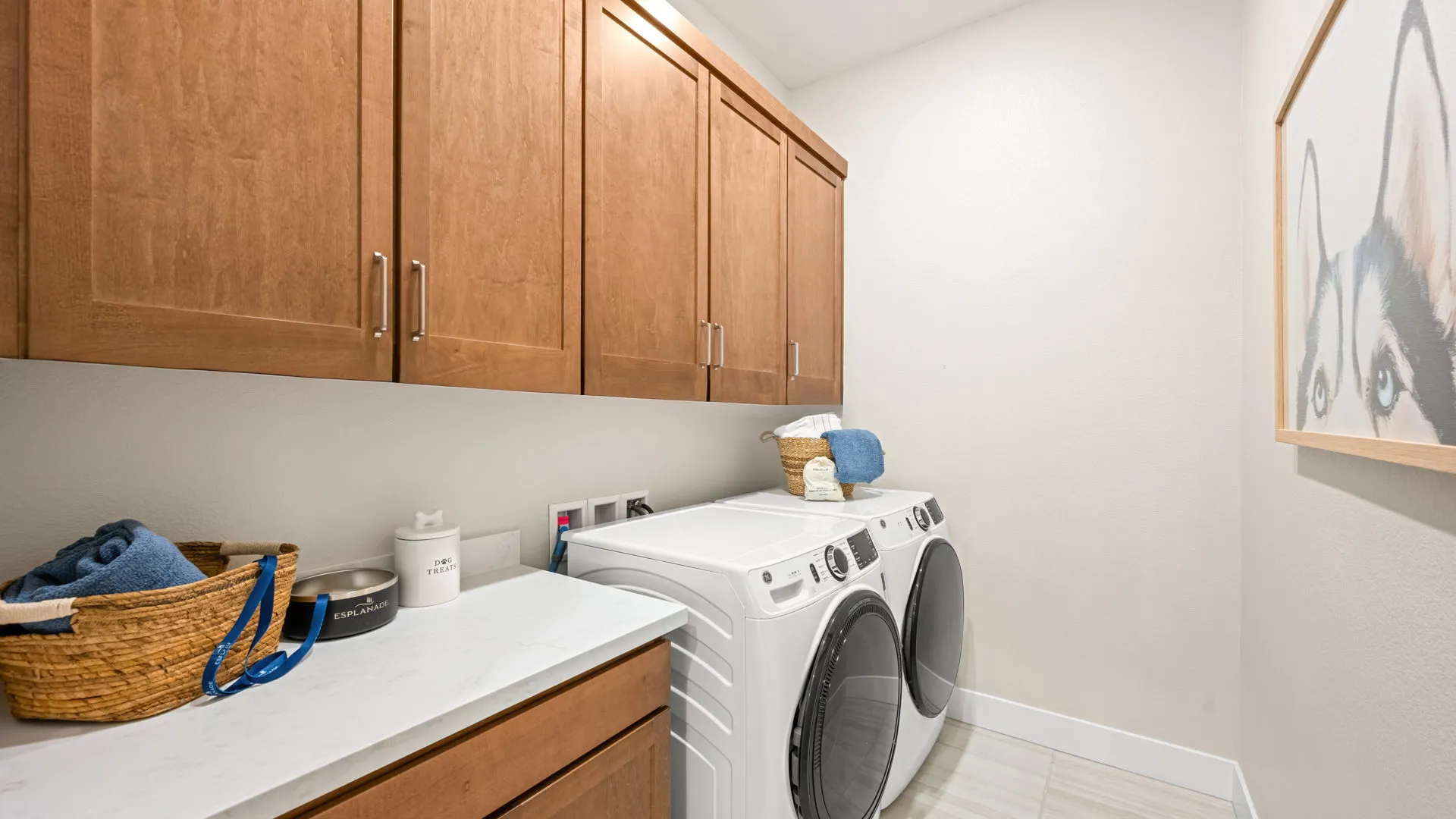 Laundry Room of Tiki Model at Esplanade at Red Rock by Taylor Morrison Laundry room with wooden cabinets, white countertop, front-loading washer and dryer, baskets with towels, and a framed picture of a dog on the wall.