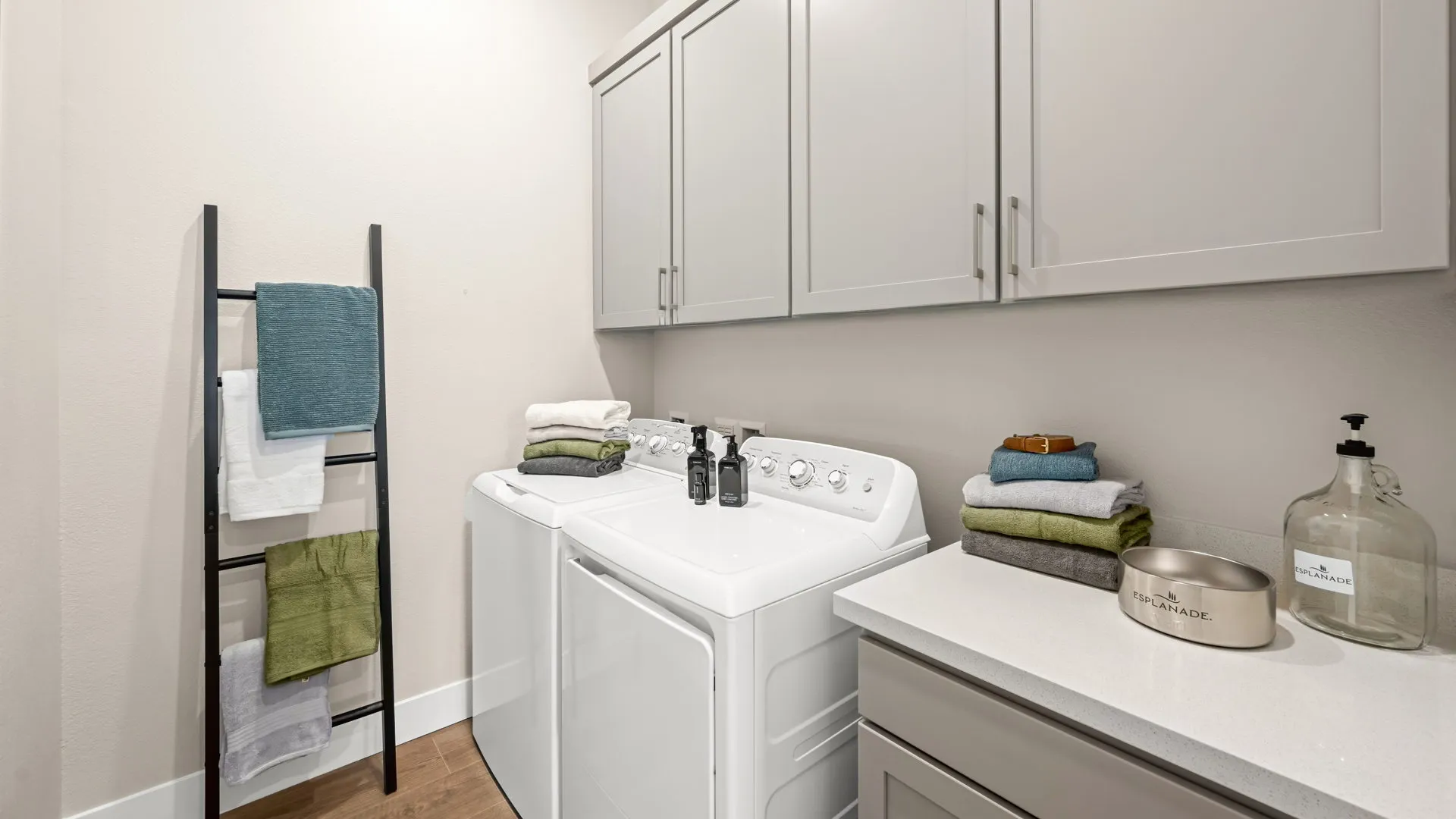 Laundry Room of Zinnia Model at Esplanade at Red Rock by Taylor Morrison A modern laundry room with a washer and dryer, white cabinets, folded towels, detergent bottles, a decorative ladder for towels, and a countertop with more towels and a bottle labeled hand soap..