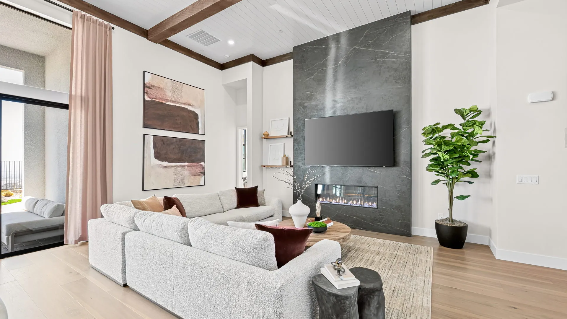 Living Room of Monroe Model at Esplanade at Red Rock by Taylor Morrison (2) Modern living room with white sectional sofa, abstract art, wall-mounted TV above a sleek fireplace, large potted plant, and neutral decor. Light wood flooring and high ceilings with exposed beams complete the space.