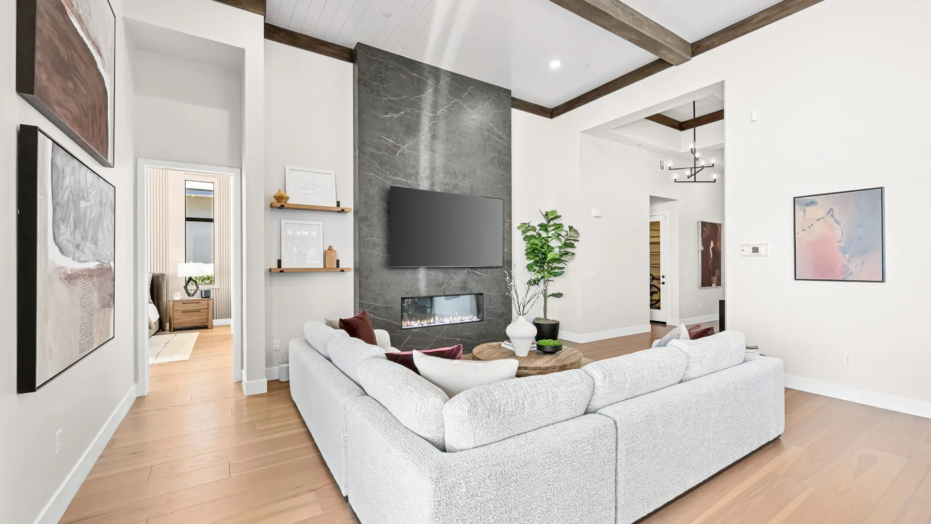 Living Room of Monroe Model at Esplanade at Red Rock by Taylor Morrison Modern living room with a light gray sectional sofa, wall-mounted TV above a fireplace, wooden shelves, large green plant, art on white walls, wood flooring, and visible entry to a bedroom.