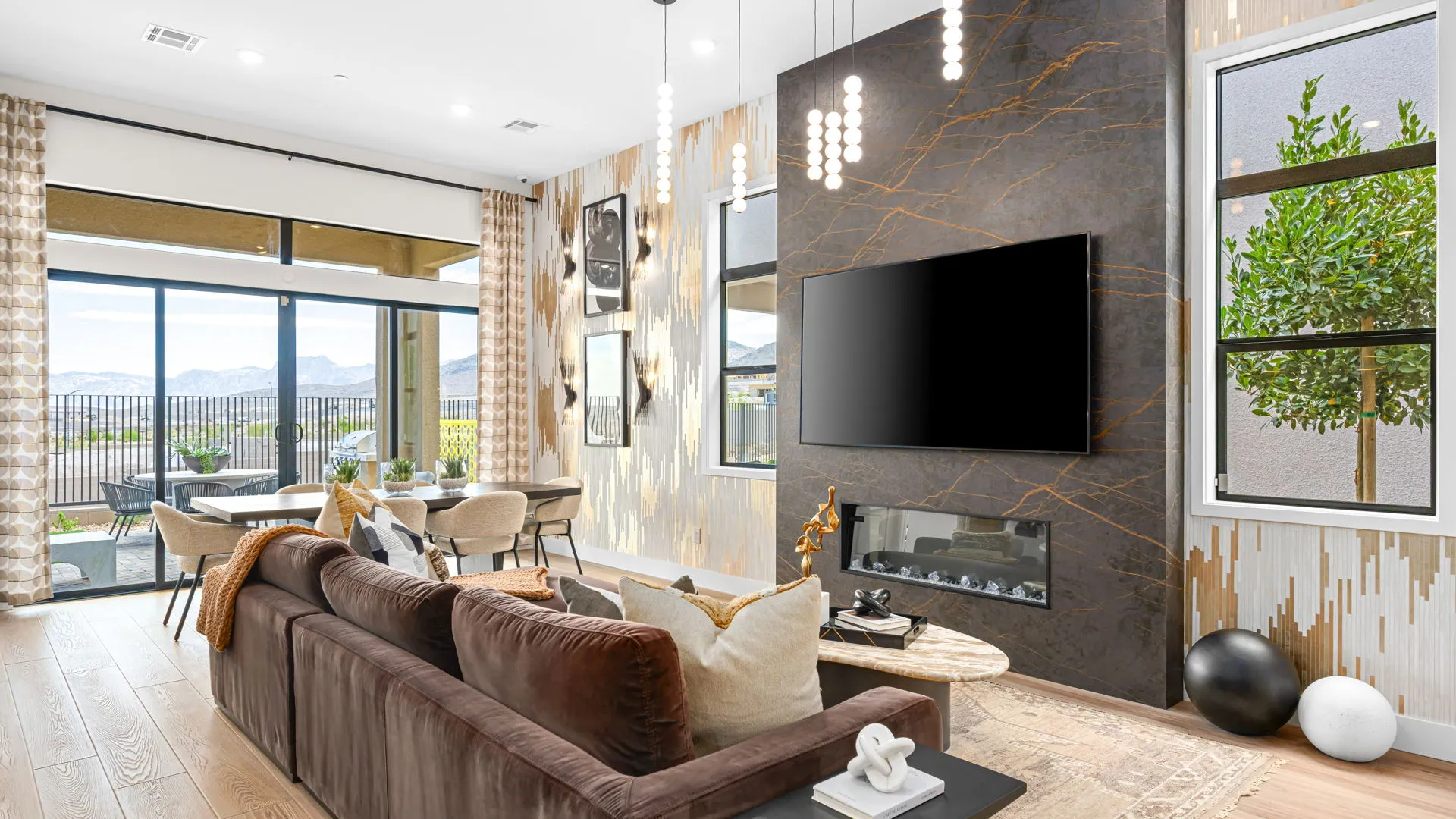 Living Room of Paloma Model at Esplanade at Red Rock by Taylor Morrison (1) Modern living room with brown sectional sofa, wall-mounted TV, linear fireplace, pendant lights, large windows, and mountain views. Dining area and neutral decor visible, with natural light filling the space.