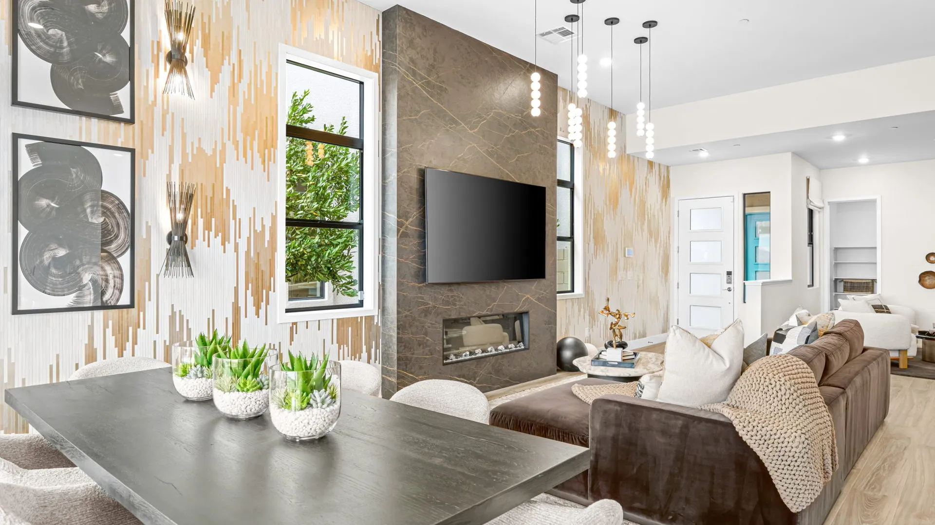 Living Room of Paloma Model at Esplanade at Red Rock by Taylor Morrison (2) Modern living room with a brown sofa, wall-mounted TV, fireplace, and contemporary decor. A dining table with potted plants sits nearby. Large windows let in natural light, brightening the neutral-toned space.