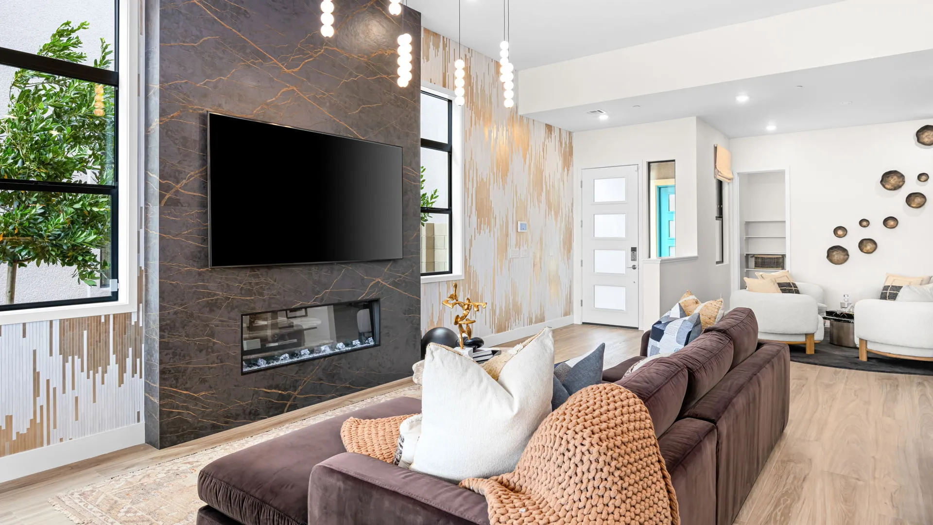 Living Room of Paloma Model at Esplanade at Red Rock by Taylor Morrison Modern living room with a brown sectional sofa, plush pillows, wall-mounted TV above a dark marble fireplace, large windows, light wood floors, and chic pendant lights. Walls have artistic patterns and decor.