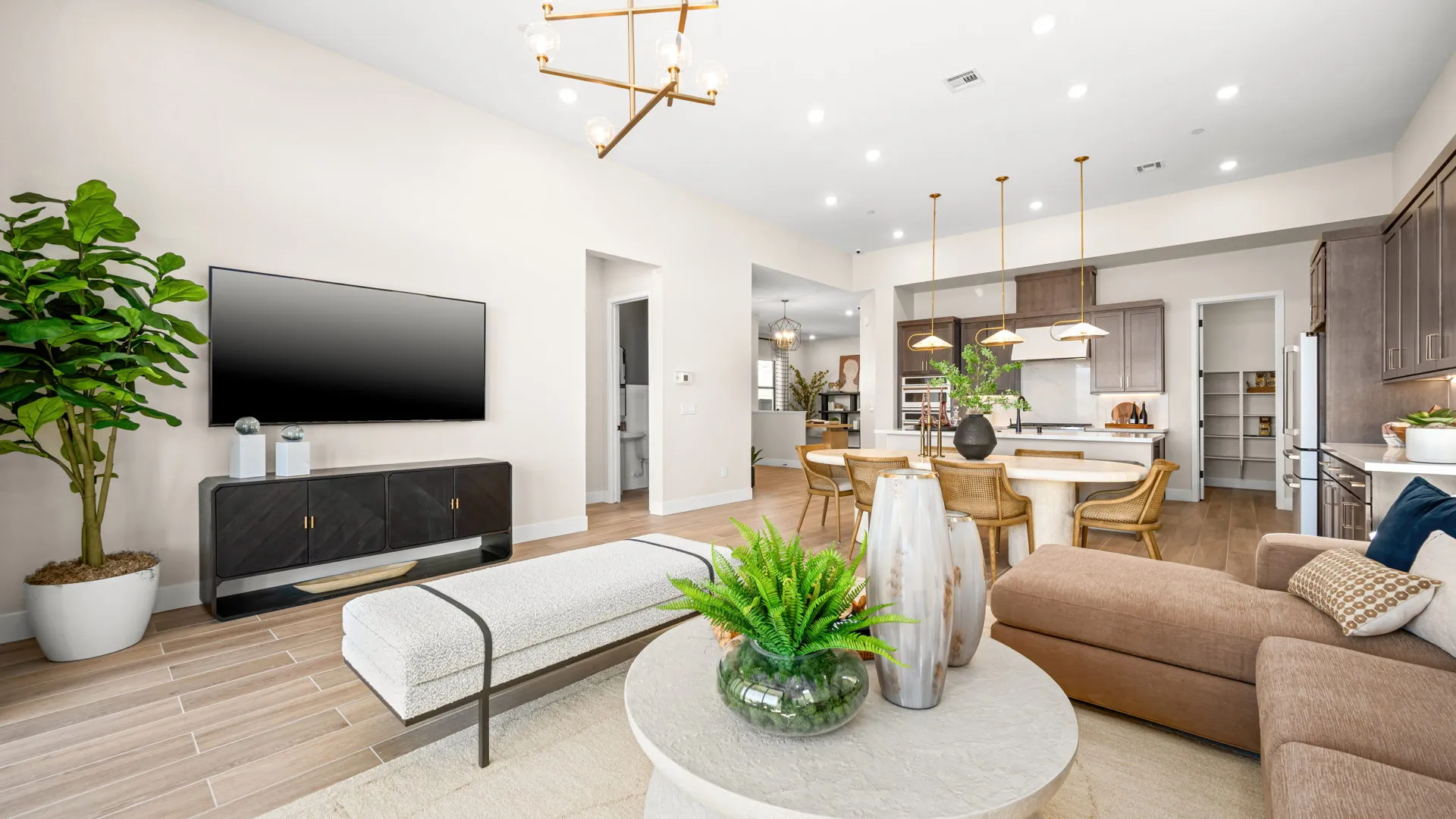 Living Room of Sangria Model at Esplanade at Red Rock by Taylor Morrison (1) Modern open-concept living room and kitchen with wooden floors, beige walls, brown sofa, TV, plants, round coffee table, and dining area with pendant lights and dark cabinets.