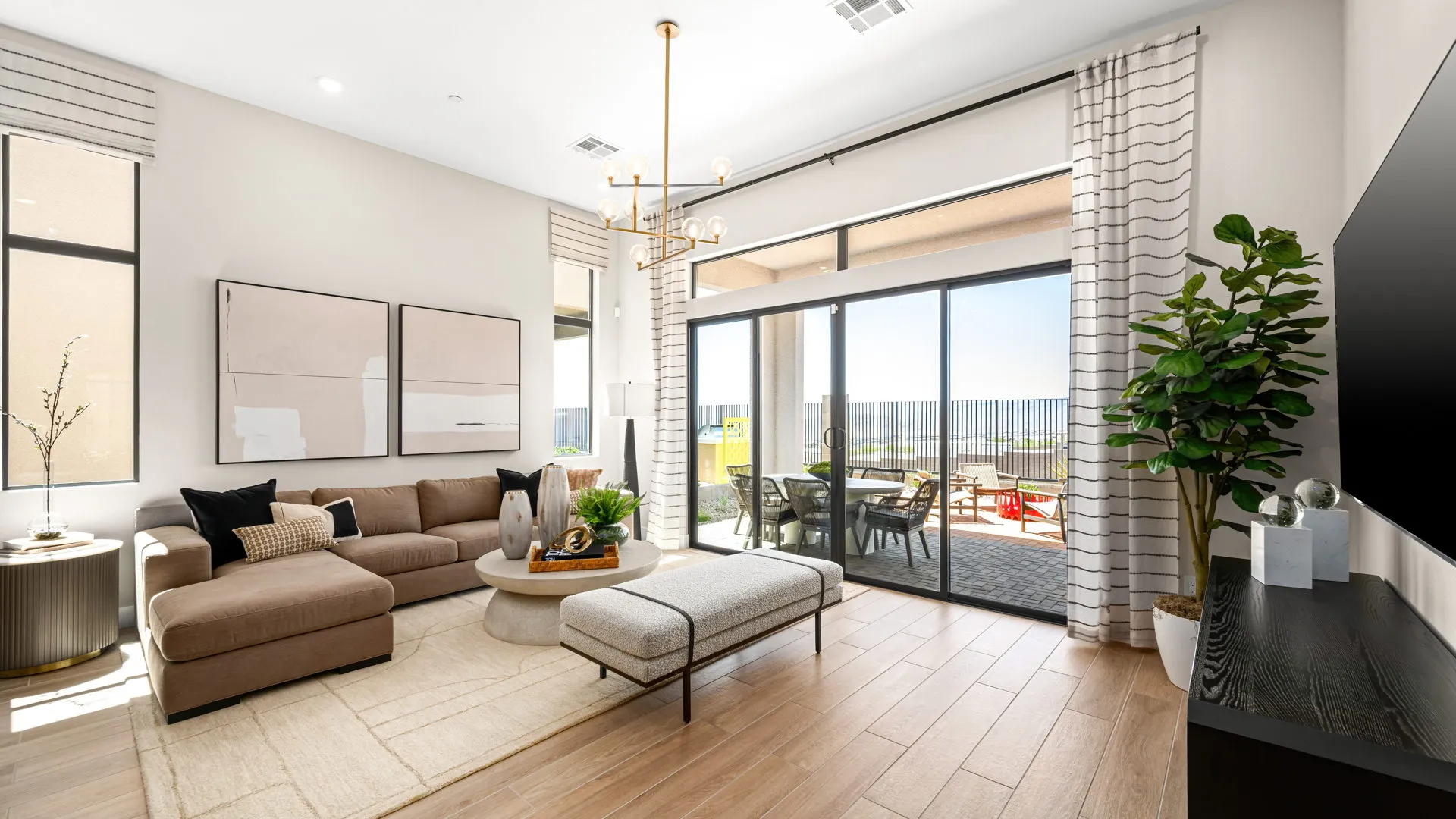 Living Room of Sangria Model at Esplanade at Red Rock by Taylor Morrison Bright living room with a tan sectional sofa, modern decor, a large potted plant, wall art, and a TV. Glass doors open to a sunny patio with a table and chairs, letting in natural light.